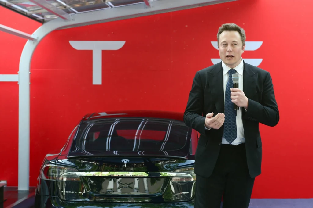 Elon Musk standing confidently in front of a sleek Tesla electric car.