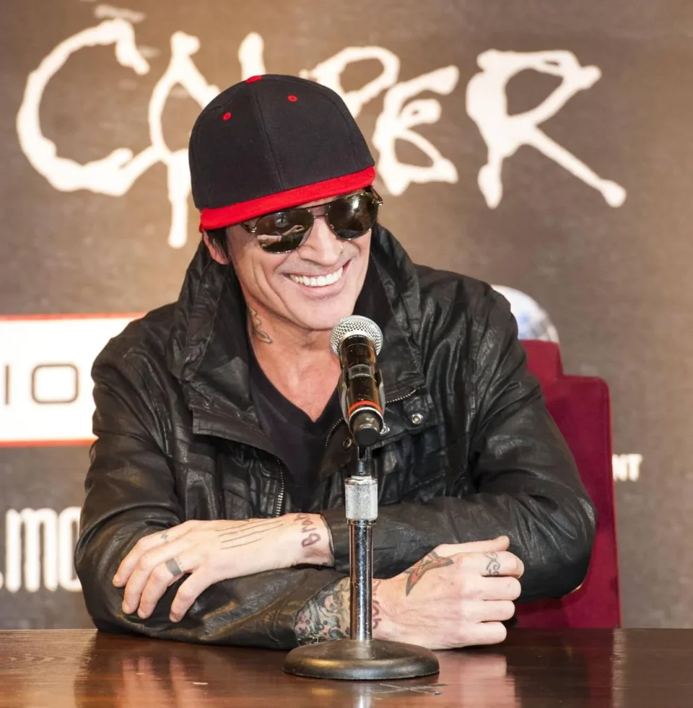 Tommy Lee sitting in an interview with a journalist, speaking passionately while wearing a black shirt and his signature tattoos visible.